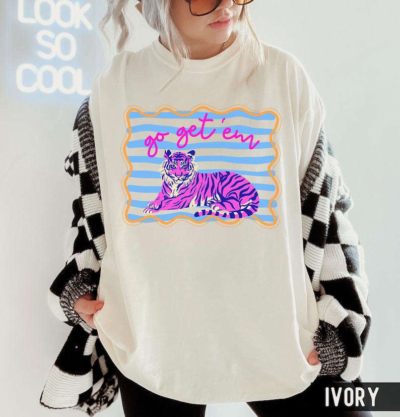 Pu&ograve; includere: T-shirt color avorio con grafica di una tigre viola a strisce blu, su sfondo a righe blu e arancioni. Il testo "go get 'em" &egrave; scritto sopra la tigre in rosa. La maglietta ha una vestibilit&agrave; ampia.