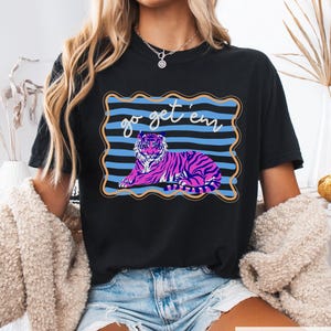 Pu&ograve; includere: T-shirt nero con un disegno di tigre rosa e viola e la scritta "go get 'em". La tigre &egrave; su uno sfondo a righe blu con un bordo dorato. La maglietta &egrave; in stile casual.