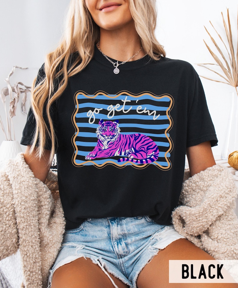 Pu&ograve; includere: T-shirt nero con un disegno di tigre rosa e viola e la scritta "go get 'em". La tigre &egrave; su uno sfondo a righe blu con un bordo dorato. La maglietta &egrave; in stile casual.