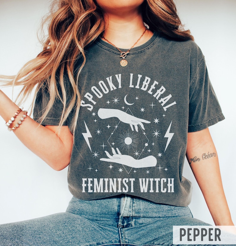 Puede incluir: Camiseta gris oscuro con el texto blanco "SPOOKY LIBERAL FEMINIST WITCH". El gr&aacute;fico incluye manos, estrellas, una luna creciente y rayos. La camiseta es de corte holgado.