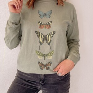 May include: A long-sleeved, sage green t-shirt with a vertical row of five different butterfly illustrations. The butterflies are in various colors, including blue, brown, yellow, green, and black.