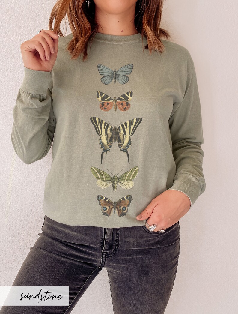 May include: A long-sleeved, sage green t-shirt with a vertical row of five different butterfly illustrations. The butterflies are in various colors, including blue, brown, yellow, green, and black.