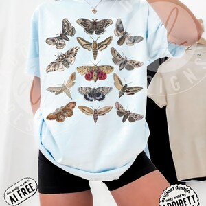 May include: A light blue t-shirt featuring a graphic print of twelve different moth species arranged in a grid. The moths are depicted in various shades of brown, tan, grey, and red, showcasing intricate wing patterns and detailed anatomical features.