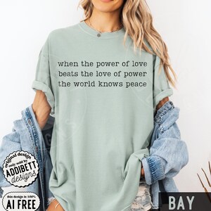 May include: A sage green short-sleeve t-shirt featuring black typewriter-style text that reads: when the power of love beats the love of power the world knows peace. The shirt is worn with a blue denim jacket draped over the shoulders.