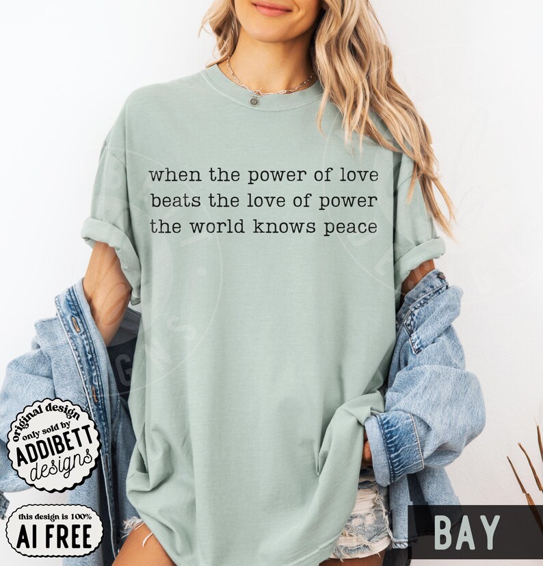 May include: A sage green short-sleeve t-shirt featuring black typewriter-style text that reads: when the power of love beats the love of power the world knows peace. The shirt is worn with a blue denim jacket draped over the shoulders.