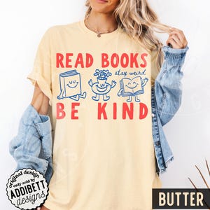 May include: A pale yellow t-shirt featuring the red text READ BOOKS BE KIND. Between the lines of text are three blue cartoon characters: a smiling book, a smiling flower vase, and an open smiling book, with the blue text stay weird.