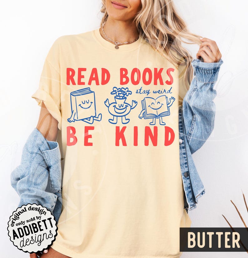 May include: A pale yellow t-shirt featuring the red text READ BOOKS BE KIND. Between the lines of text are three blue cartoon characters: a smiling book, a smiling flower vase, and an open smiling book, with the blue text stay weird.