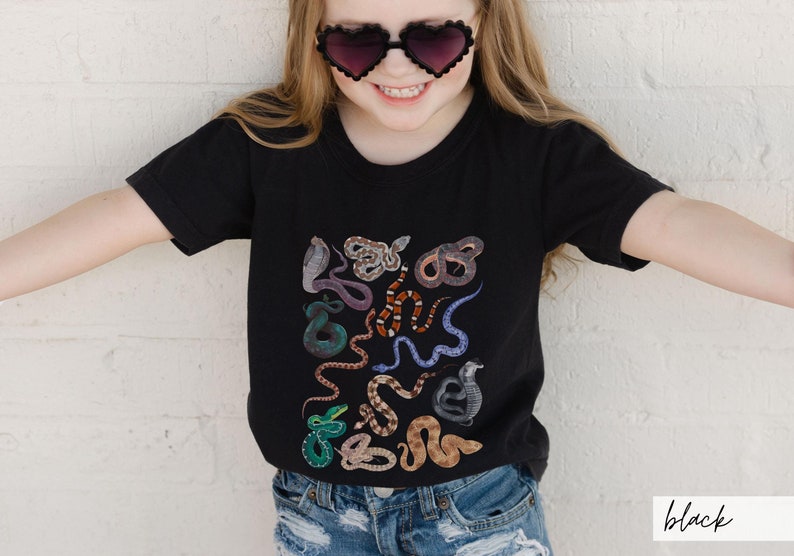 Pode incluir: Uma camiseta preta com um design de estampa de cobras coloridas. A camiseta est&aacute; sendo usada por uma crian&ccedil;a que usa &oacute;culos de sol em forma de cora&ccedil;&atilde;o.