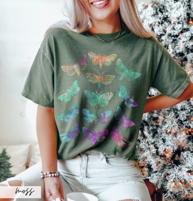 May include: A green t-shirt featuring a colourful moth design. The moths are arranged in a circular pattern and have iridescent wings.