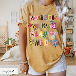 May include: A mustard yellow t-shirt with a graphic print that reads "Read Books Be Kind Stay Weird" in colorful, cut-out letters.