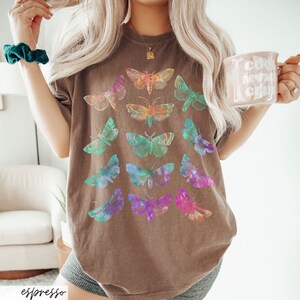 May include: An oversized brown t-shirt featuring a colourful butterfly graphic print. The butterflies are arranged in a pattern and are iridescent.