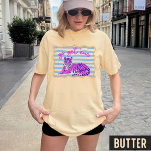 Pu&ograve; includere: T-shirt color burro con grafica di tigre e la scritta "go get 'em". La tigre &egrave; rosa e viola, su sfondo a righe blu. La maglietta ha una vestibilit&agrave; ampia e oversize.