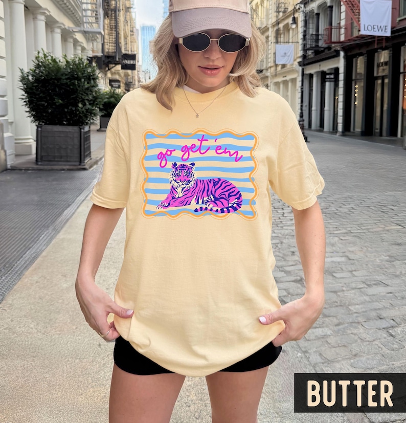 Pu&ograve; includere: T-shirt color burro con grafica di tigre e la scritta "go get 'em". La tigre &egrave; rosa e viola, su sfondo a righe blu. La maglietta ha una vestibilit&agrave; ampia e oversize.