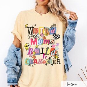 May include: Butter-colored t-shirt with the text "Weird Moms Build Character" in a colorful, playful design. The graphic includes flowers, stars, and a checkered heart. The shirt has short sleeves and a relaxed fit.