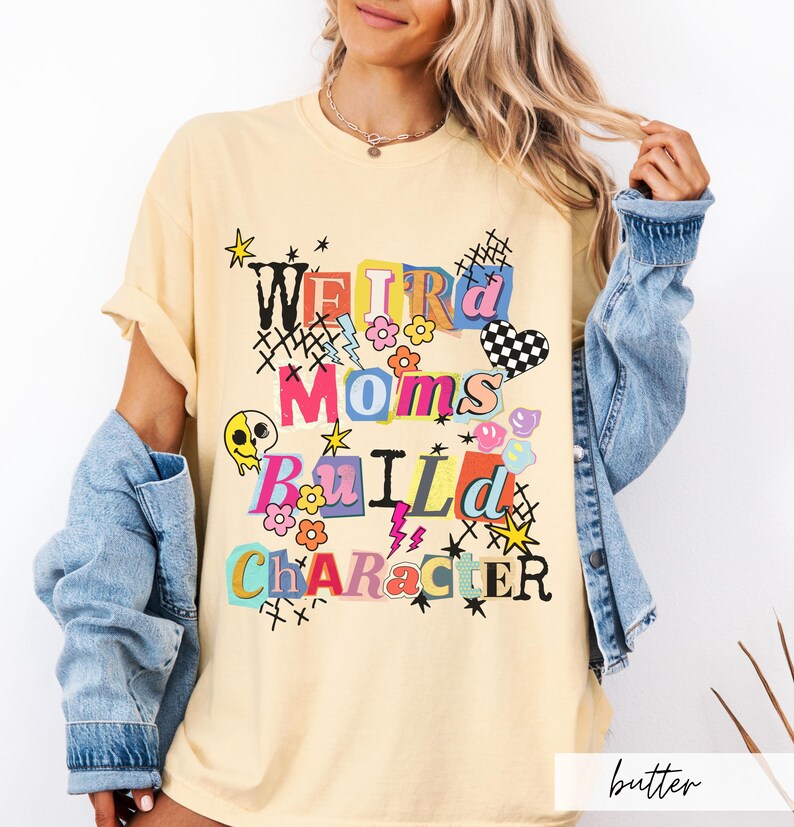 May include: Butter-colored t-shirt with the text "Weird Moms Build Character" in a colorful, playful design. The graphic includes flowers, stars, and a checkered heart. The shirt has short sleeves and a relaxed fit.