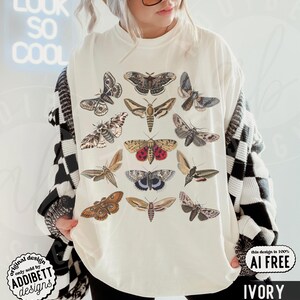 May include: An ivory t-shirt featuring a graphic print of various moth species in shades of brown, tan, gray, and red. The moths are arranged in a grid-like pattern across the front of the shirt. The garment has a crew neckline and short sleeves.