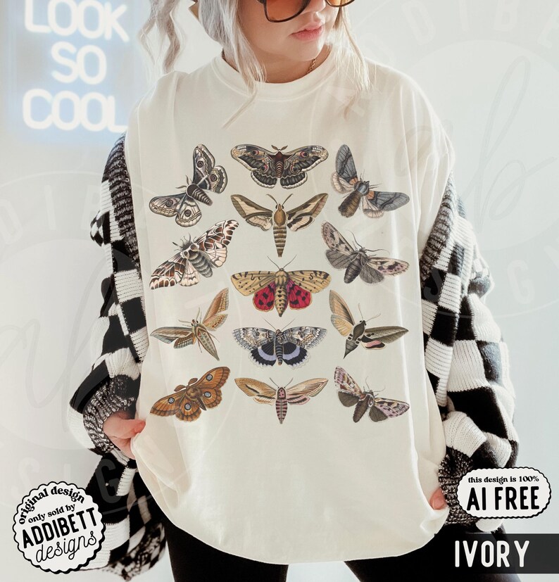 May include: An ivory t-shirt featuring a graphic print of various moth species in shades of brown, tan, gray, and red. The moths are arranged in a grid-like pattern across the front of the shirt. The garment has a crew neckline and short sleeves.