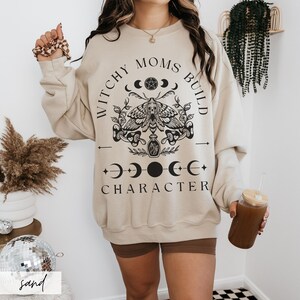 K&ouml;nnte beinhalten: Sandfarbenes Sweatshirt mit dem schwarzen Text "Witchy Moms Build Character". Die Grafik zeigt eine Motte, Halbmonde und andere mystische Symbole. Das Sweatshirt hat eine lockere Passform.