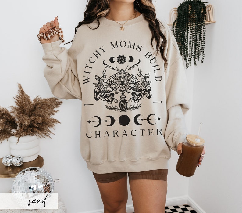 K&ouml;nnte beinhalten: Sandfarbenes Sweatshirt mit dem schwarzen Text "Witchy Moms Build Character". Die Grafik zeigt eine Motte, Halbmonde und andere mystische Symbole. Das Sweatshirt hat eine lockere Passform.
