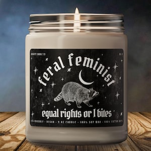 Può includere: Una candela bianca in barattolo con uno stoppino acceso. L'etichetta recita "feral feminist" con un'illustrazione di procione e il testo "equal rights or I bites". La candela è da 255 grammi ed è fatta con cera di soia.