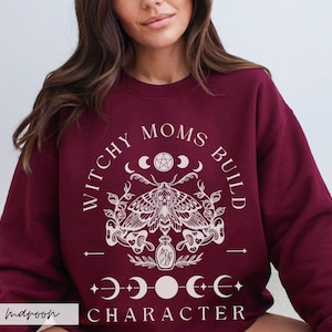 K&ouml;nnte beinhalten: Burgunderfarbenes Sweatshirt mit dem wei&szlig;en Text "WITCHY MOMS BUILD CHARACTER". Das Design umfasst eine Motte, Halbmonde und ein Pentagramm. Das Sweatshirt hat einen Rundhalsausschnitt.