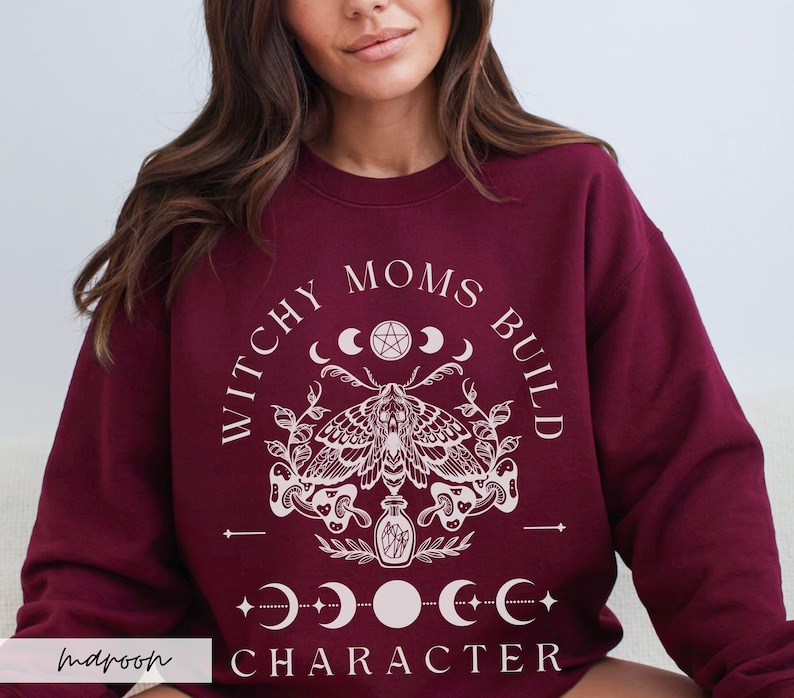 K&ouml;nnte beinhalten: Burgunderfarbenes Sweatshirt mit dem wei&szlig;en Text "WITCHY MOMS BUILD CHARACTER". Das Design umfasst eine Motte, Halbmonde und ein Pentagramm. Das Sweatshirt hat einen Rundhalsausschnitt.