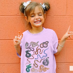 Pode incluir: Uma pessoa jovem vestindo uma camiseta lil&aacute;s clara com uma estampa de cobra colorida. A camiseta tem gola redonda e mangas curtas. A pessoa est&aacute; sorrindo e tem o cabelo puxado para tr&aacute;s com dois la&ccedil;os brancos.