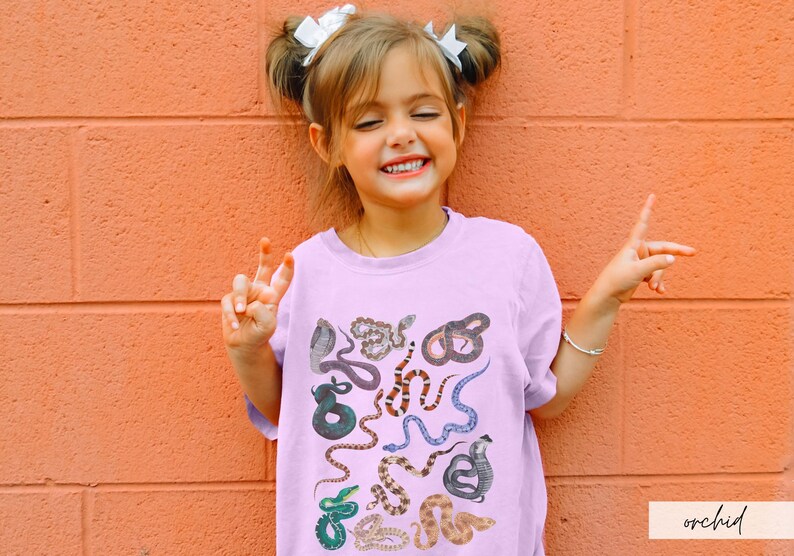 Pode incluir: Uma pessoa jovem vestindo uma camiseta lil&aacute;s clara com uma estampa de cobra colorida. A camiseta tem gola redonda e mangas curtas. A pessoa est&aacute; sorrindo e tem o cabelo puxado para tr&aacute;s com dois la&ccedil;os brancos.