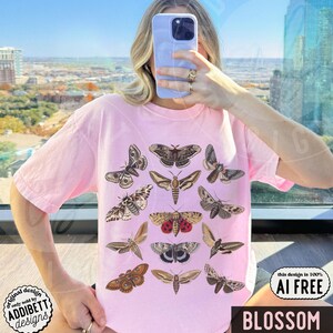 May include: A light pink short-sleeve t-shirt featuring a graphic print of twelve different moth species in various shades of brown, tan, and red. The moths are arranged in a grid-like pattern across the center of the chest area of the garment.