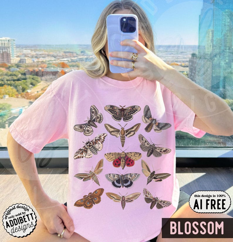 May include: A light pink short-sleeve t-shirt featuring a graphic print of twelve different moth species in various shades of brown, tan, and red. The moths are arranged in a grid-like pattern across the center of the chest area of the garment.