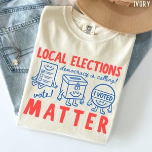 Op de afbeelding: Ivoor kleurig t-shirt met de rode woorden "LOCAL ELECTIONS MATTER" en de blauwe zin "democracy is calling!". Het shirt heeft cartoon illustraties van een stembiljet, een stembus en een "I Voted" knop.