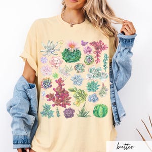 Puede incluir: Una camiseta amarilla con un estampado acuarela colorido de diferentes suculentas. El estampado presenta una variedad de formas, tama&ntilde;os y colores de suculentas.