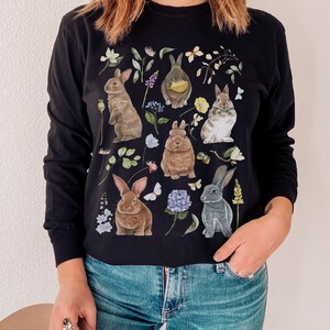 May include: Black long-sleeved shirt with a graphic print of multiple rabbits and flowers. The rabbits are brown, gray, and white.