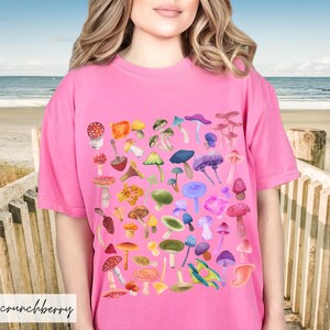 K&ouml;nnte beinhalten: Pinkes T-Shirt mit einem bunten Pilzdruck. Die Pilze sind in einem quadratischen Muster angeordnet und in verschiedenen Schattierungen von Rot, Orange, Gelb, Gr&uuml;n, Blau und Lila.
