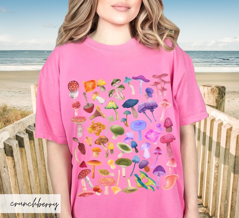 K&ouml;nnte beinhalten: Pinkes T-Shirt mit einem bunten Pilzdruck. Die Pilze sind in einem quadratischen Muster angeordnet und in verschiedenen Schattierungen von Rot, Orange, Gelb, Gr&uuml;n, Blau und Lila.