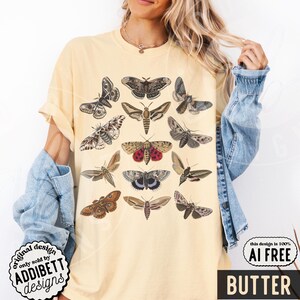 May include: A pale yellow short-sleeve t-shirt featuring a graphic print of various moth species in shades of brown, gray, and red. The design displays twelve distinct moth illustrations arranged in a grid pattern across the front of the garment.