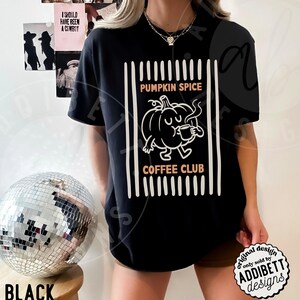 May include: A black short-sleeve t-shirt featuring a white graphic of a cartoon pumpkin drinking from a mug. The text PUMPKIN SPICE COFFEE CLUB is printed in orange above and below the pumpkin, framed by vertical white lines on both sides.