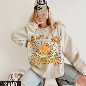 Pu&ograve; includere: Felpa color sabbia con la scritta "STUFF ME Daddy" e una grafica di tacchino dei cartoni animati. Il tacchino &egrave; su un piatto con fiori. La persona indossa un berretto da baseball bianco con la scritta "KARMA".