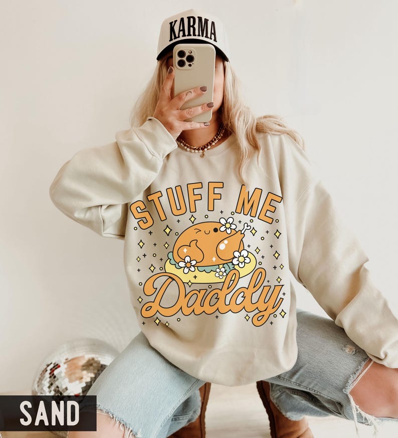 Pu&ograve; includere: Felpa color sabbia con la scritta "STUFF ME Daddy" e una grafica di tacchino dei cartoni animati. Il tacchino &egrave; su un piatto con fiori. La persona indossa un berretto da baseball bianco con la scritta "KARMA".