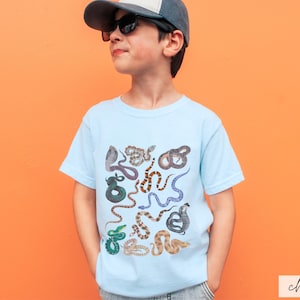 Pode incluir: Uma camiseta azul claro com uma estampa colorida de v&aacute;rias cobras. A camiseta est&aacute; sendo usada por uma crian&ccedil;a que est&aacute; usando um bon&eacute; de beisebol preto e &oacute;culos de sol.