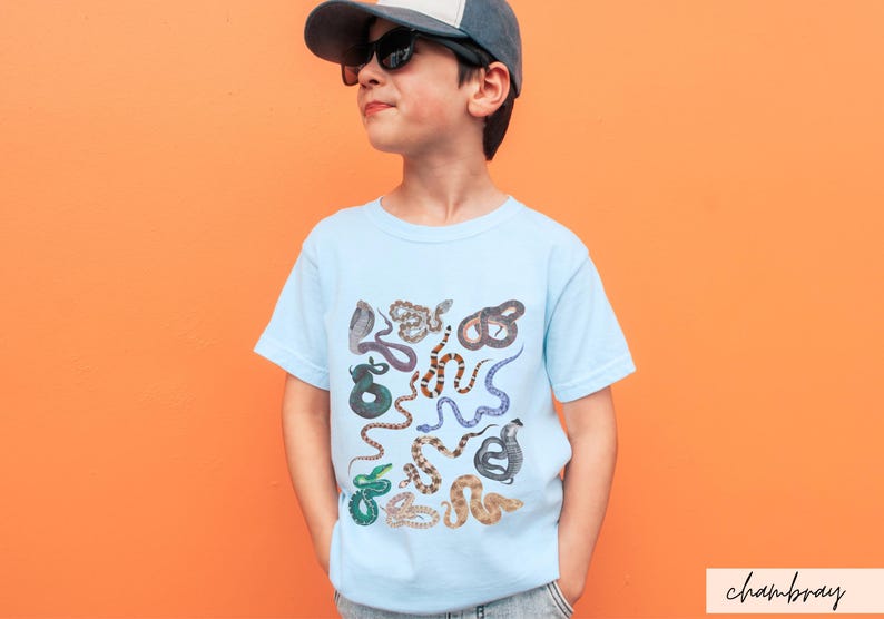 Pode incluir: Uma camiseta azul claro com uma estampa colorida de v&aacute;rias cobras. A camiseta est&aacute; sendo usada por uma crian&ccedil;a que est&aacute; usando um bon&eacute; de beisebol preto e &oacute;culos de sol.