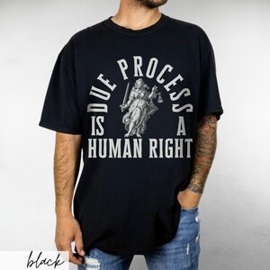 May include: Black t-shirt with a white graphic print of a woman holding scales of justice. The text reads "Due Process Is A Human Right".