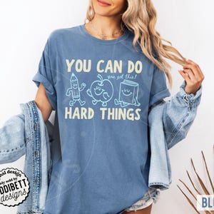 K&ouml;nnte beinhalten: Blaues Jeans-T-Shirt mit dem Text "YOU CAN DO HARD THINGS" und dem Satz "you got this!". Das Design umfasst Cartoon-Illustrationen eines Bleistifts, Apfels und Buches. Das T-Shirt hat eine lockere Passform und ist ein Freizeitkleidungsst&uuml;ck.