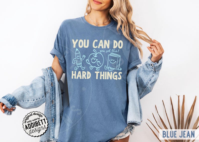 K&ouml;nnte beinhalten: Blaues Jeans-T-Shirt mit dem Text "YOU CAN DO HARD THINGS" und dem Satz "you got this!". Das Design umfasst Cartoon-Illustrationen eines Bleistifts, Apfels und Buches. Das T-Shirt hat eine lockere Passform und ist ein Freizeitkleidungsst&uuml;ck.