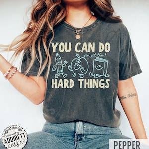 K&ouml;nnte beinhalten: Dunkelgraues T-Shirt mit dem Text "YOU CAN DO HARD THINGS" und Cartoon-Illustrationen eines Bleistifts, Apfels und Buches. Das Shirt hat auch den Text "you got this!" und das Wort "PEPPER" unten.