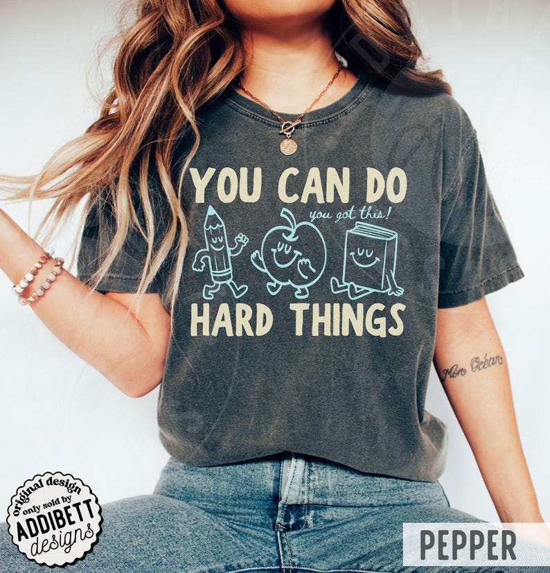 K&ouml;nnte beinhalten: Dunkelgraues T-Shirt mit dem Text "YOU CAN DO HARD THINGS" und Cartoon-Illustrationen eines Bleistifts, Apfels und Buches. Das Shirt hat auch den Text "you got this!" und das Wort "PEPPER" unten.