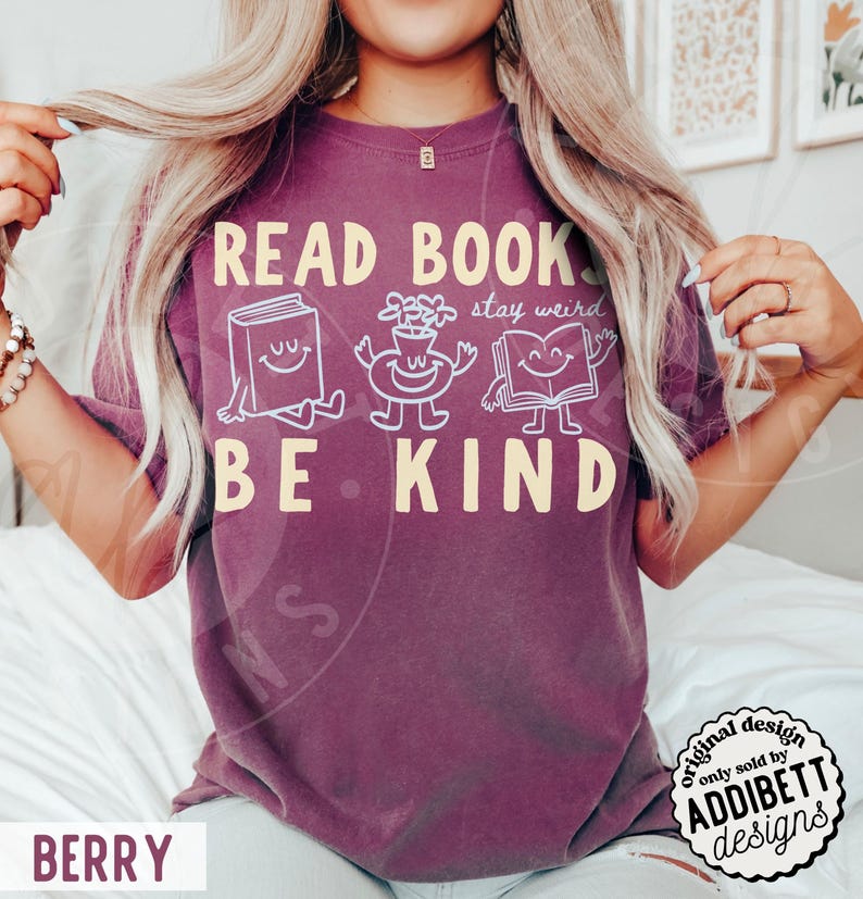 May include: A berry-colored short-sleeve t-shirt featuring the yellow text READ BOOKS BE KIND and white line-art illustrations of a smiling book, a flower vase with a face, and an open book with arms, plus the cursive text stay weird.
