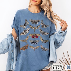 May include: A blue denim-colored t-shirt featuring a graphic print of various moth species in shades of brown, tan, grey, and red. The shirt has short sleeves and a crew neckline, showcasing a detailed collection of insect illustrations arranged in rows.