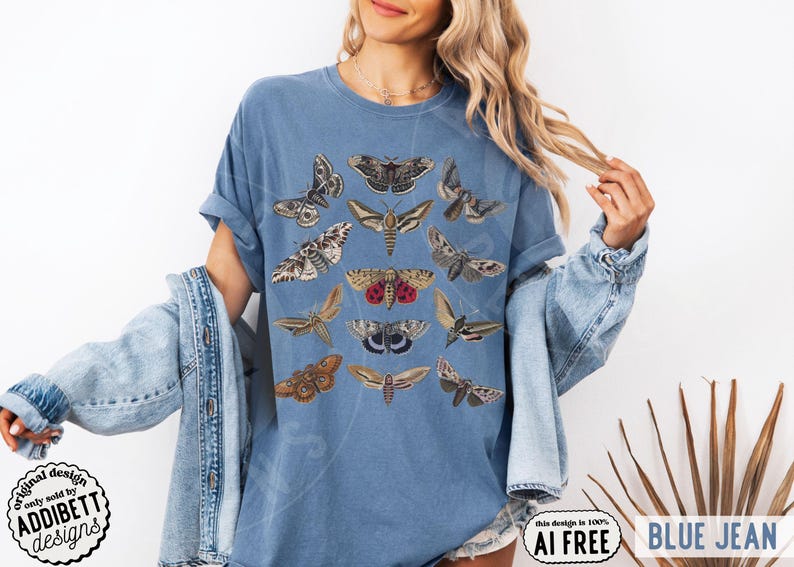 May include: A blue denim-colored t-shirt featuring a graphic print of various moth species in shades of brown, tan, grey, and red. The shirt has short sleeves and a crew neckline, showcasing a detailed collection of insect illustrations arranged in rows.