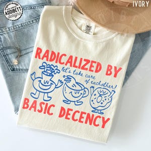 Pode incluir: Camiseta cor marfim com o texto vermelho "RADICALIZED BY BASIC DECENCY" e ilustrações azuis de desenhos animados. A camiseta, da coleção Comfort Colors, é exibida com jeans azuis e um colar. Feita de um material macio.